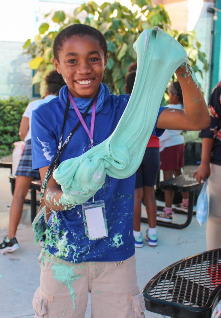 Student smiles while he stretches green slime during science club.
